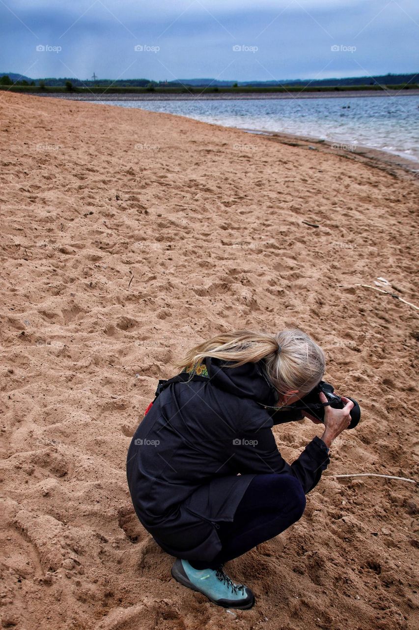 A blonde woman with ponytail crouches on a sandy beach in cool weather and an overcast sky, holding a digital camera in her hand 