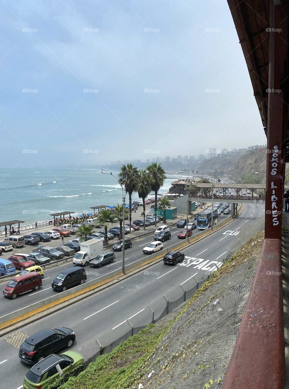 Cars on road and cars parked along ocean coastline and ocean with palm trees and skyline in background.