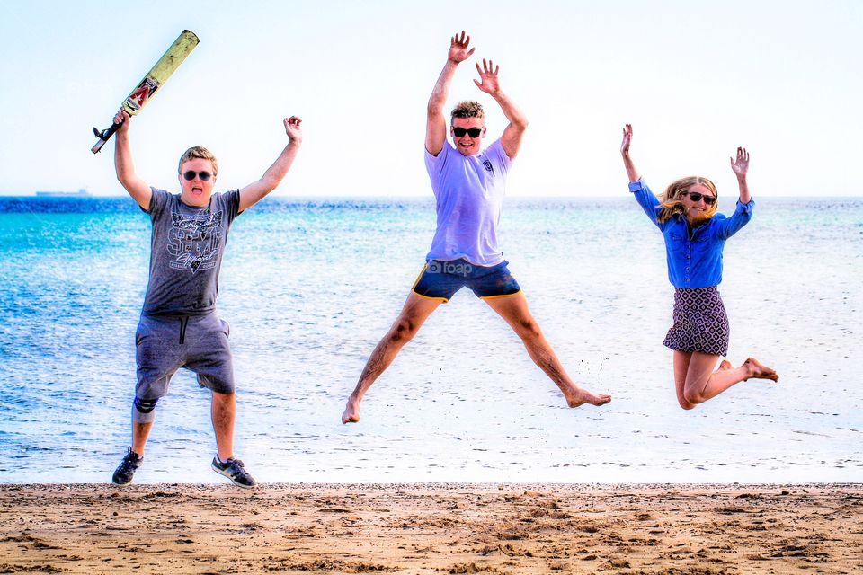 Three teenagers jumping in unison on a sandy beach with the ocean behind them.