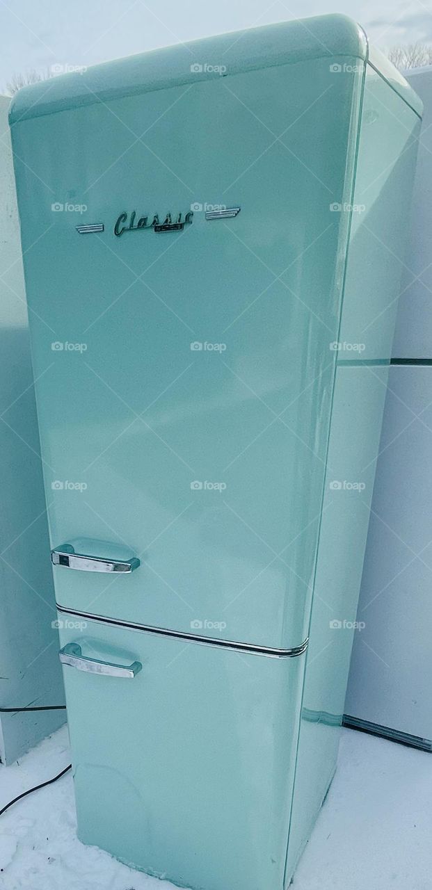 A mint green Classic Retro Bottom Freezer Refrigerator stands abandoned at the local transfer station, adding a pop of color to the dull surroundings. The fridge, with its vintage appeal, has chrome handles and rounded edges that evoke nostalgia.