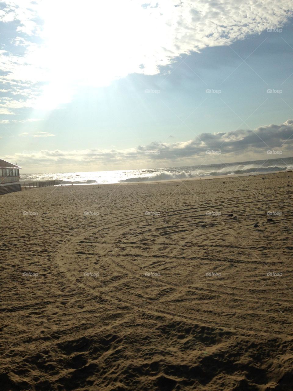 Tracks form a circular pattern in the sand in this photo of the beach taken from the boardwalk in Point Pleasant Beach, NJ. The wide stretch of sand is the focus of the photo, highlighted by ocean waves, clouds, and a streak of sun.