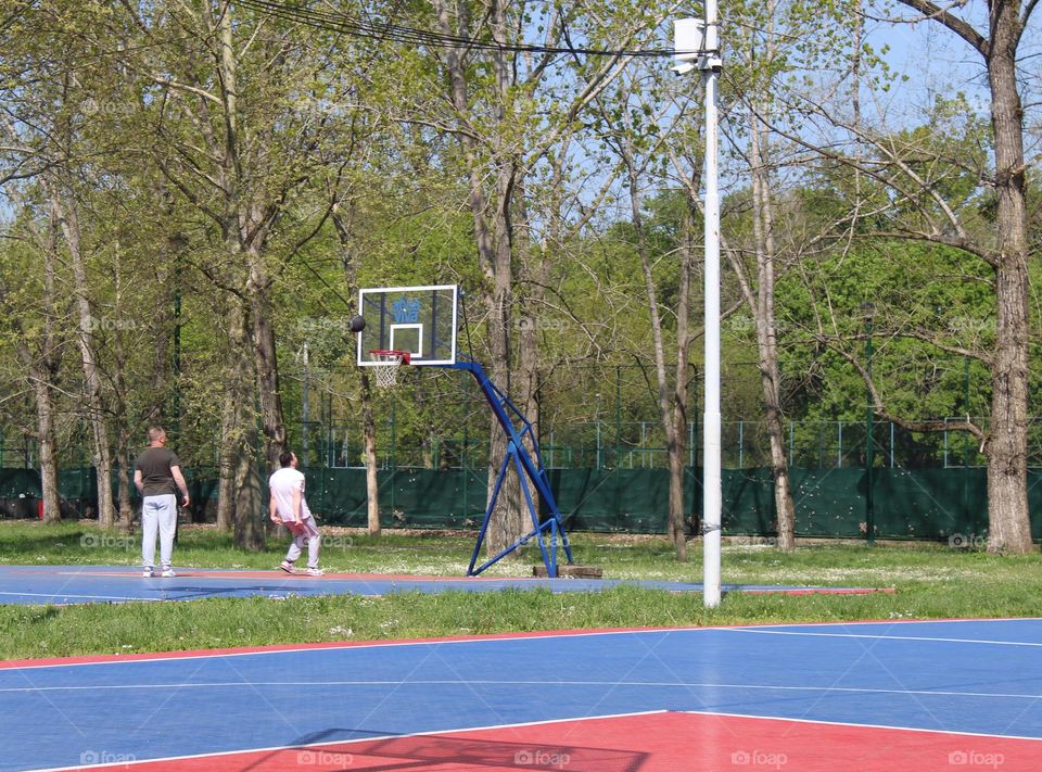 Nice spring day for recreation.  Two grown men are playing basketball in a public park