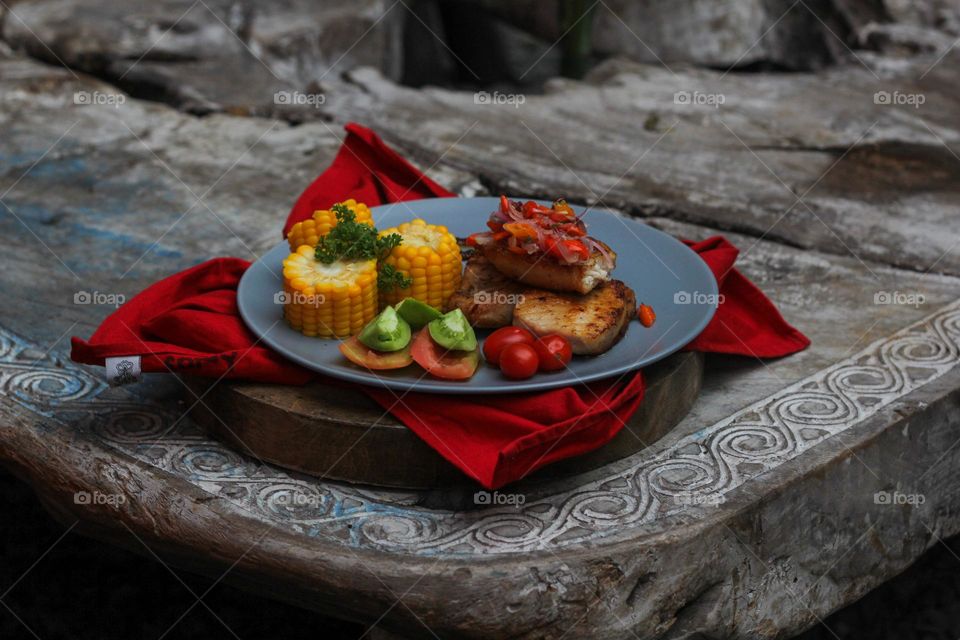 Tuna steak cooked with corn garnished with cherries, sliced tomatoes and lemon topped with sliced hot chili peppers on a baby blue melamine plate lined with red cloth and thick wood. Restaurant food.