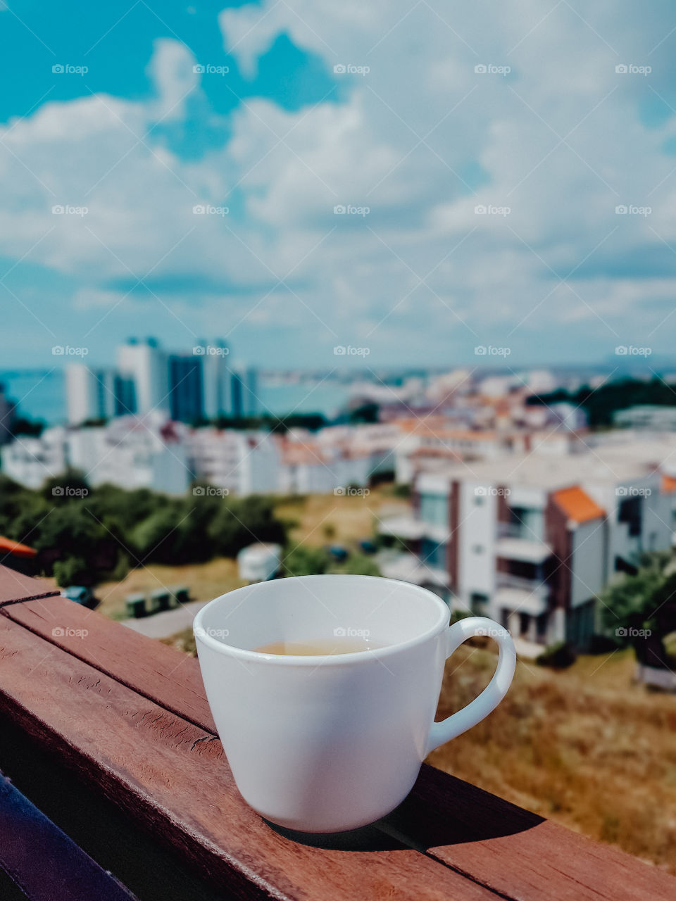 Selective focus to cup with a drink on the balcony overlooking to blurry city by the sea and cloudy sky.