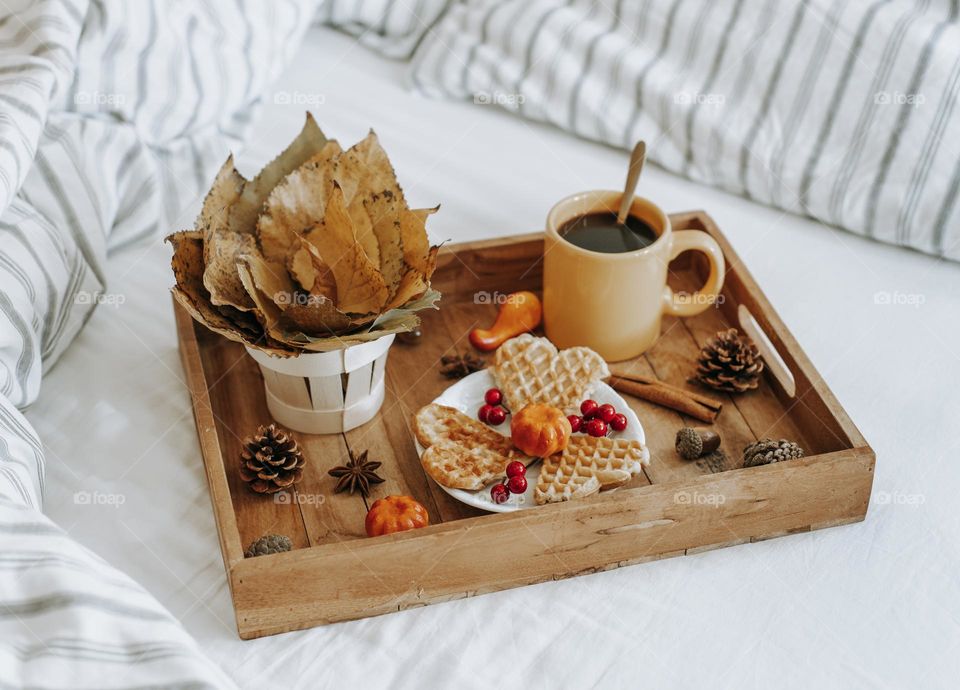 Wooden tray with hot coffee, fresh Belgian waffles and autumn leaves in bed early in the morning, side view close-up.