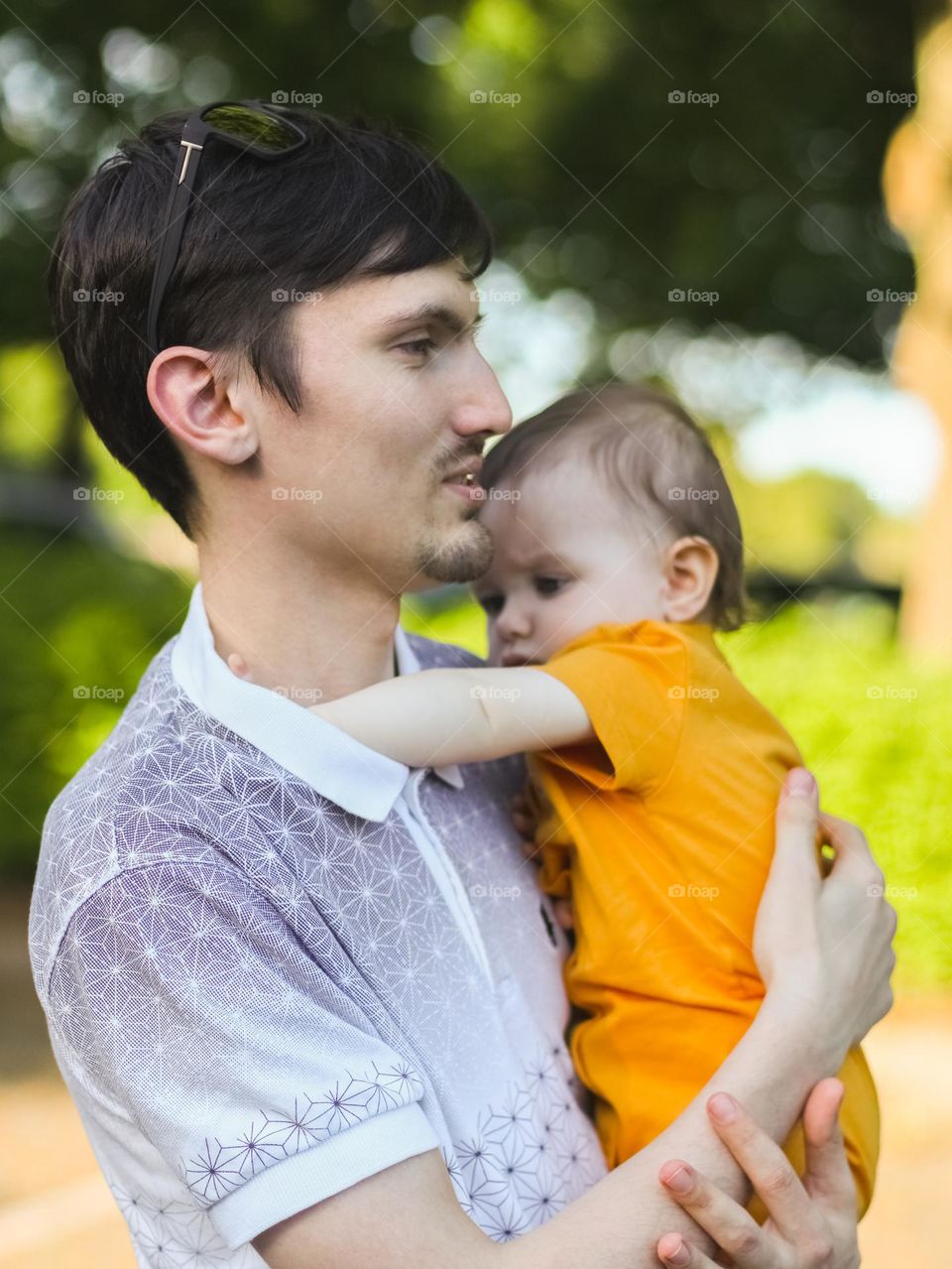 A handsome young Caucasian father with short brown hair holds his little daughter in his arms, who climbs his t-shirt with his hand while playing with him, side view close-up. Concept fathers, outdoor play, outdoor walk, young fathers, modern lifesty