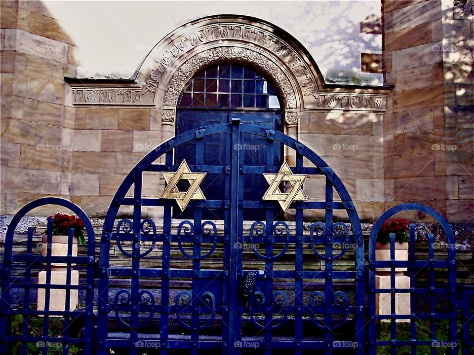 Exterior of a synagogue in a Bavarian town w two golden stars of David on the arched metal entrance gates. The large arched center window has an inscription in flaming Hebrew letters on the ornamented stone facade over it. 2021. Hypnotic Productions🏐