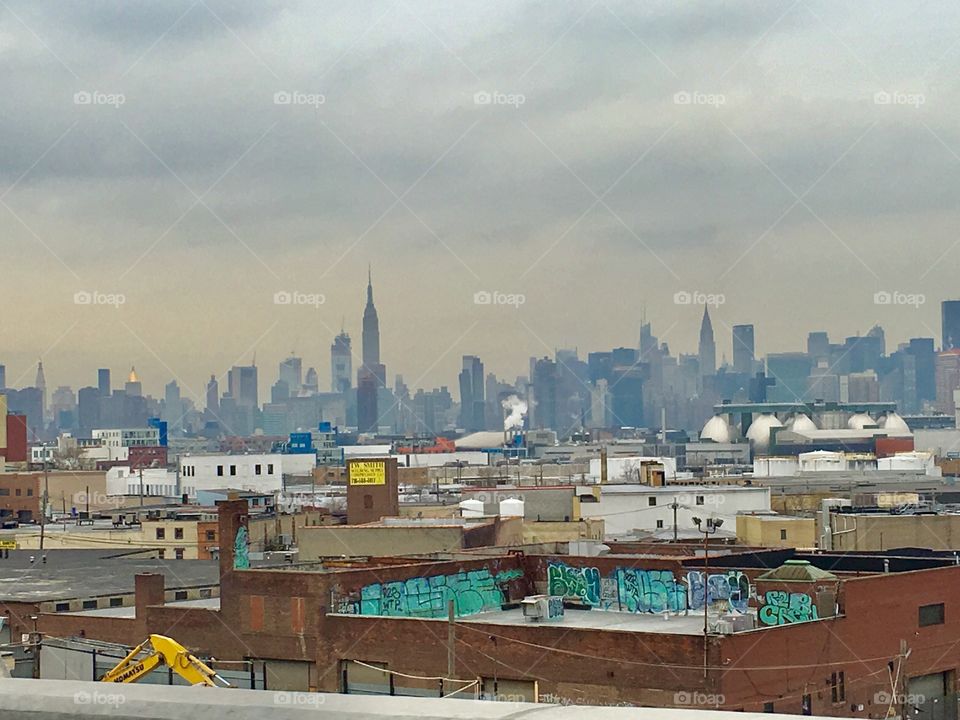 An urban view at twilight time with Long Island City, Queens, NY in the foreground underneath an overcast silvery sky and the faint blue silhouette of the Manhattan skyline in the far distance. 2017. Hypnotic Productions