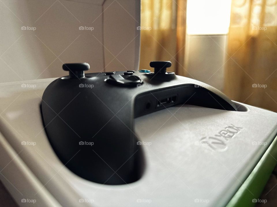 Black Xbox controller inside of its box just opened, while the natural light enters through the window.