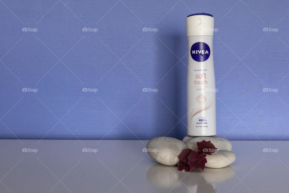 One bottle of anti-transpirant brand nivea stands on three stones with burgundy flower leaves on a white chest of drawers in the bedroom on the right against a lilac blue wall with copy space on the left, close-up side view.