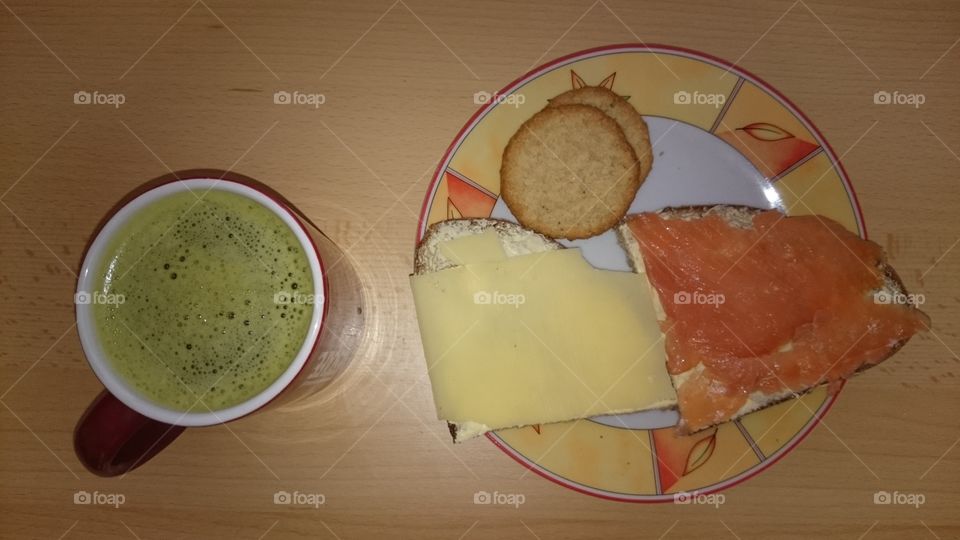 A meal consisting of a cup of matcha green tea, two slices of bread with butter and as topping salmon and cheese respectively, two swedish oat cookies