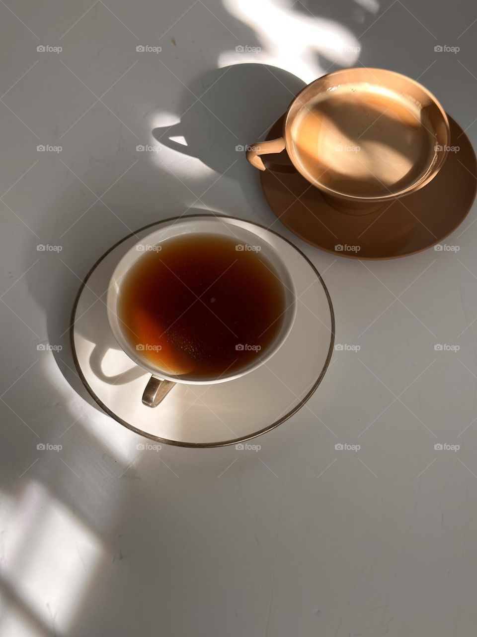 A cup of tea in a white with gold rimmed porcelain cup with saucer and a cup of coffee with cream in a brown plastic cup and saucer in afternoon light