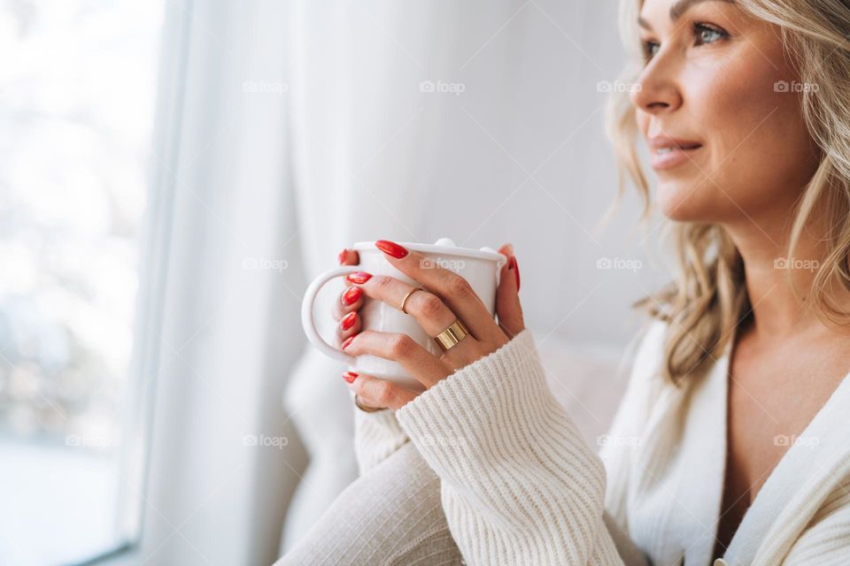 Young woman in white cardigan with cup of cocoa looking at window with winter landscape at home