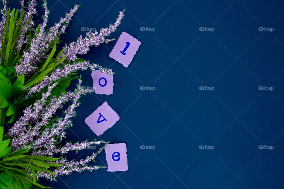 Love is all around! Lavender flowers and torn squares of lavender paper with the letters spelling “l-o-v-e” stamped in dark blue ink on a dark blue background 