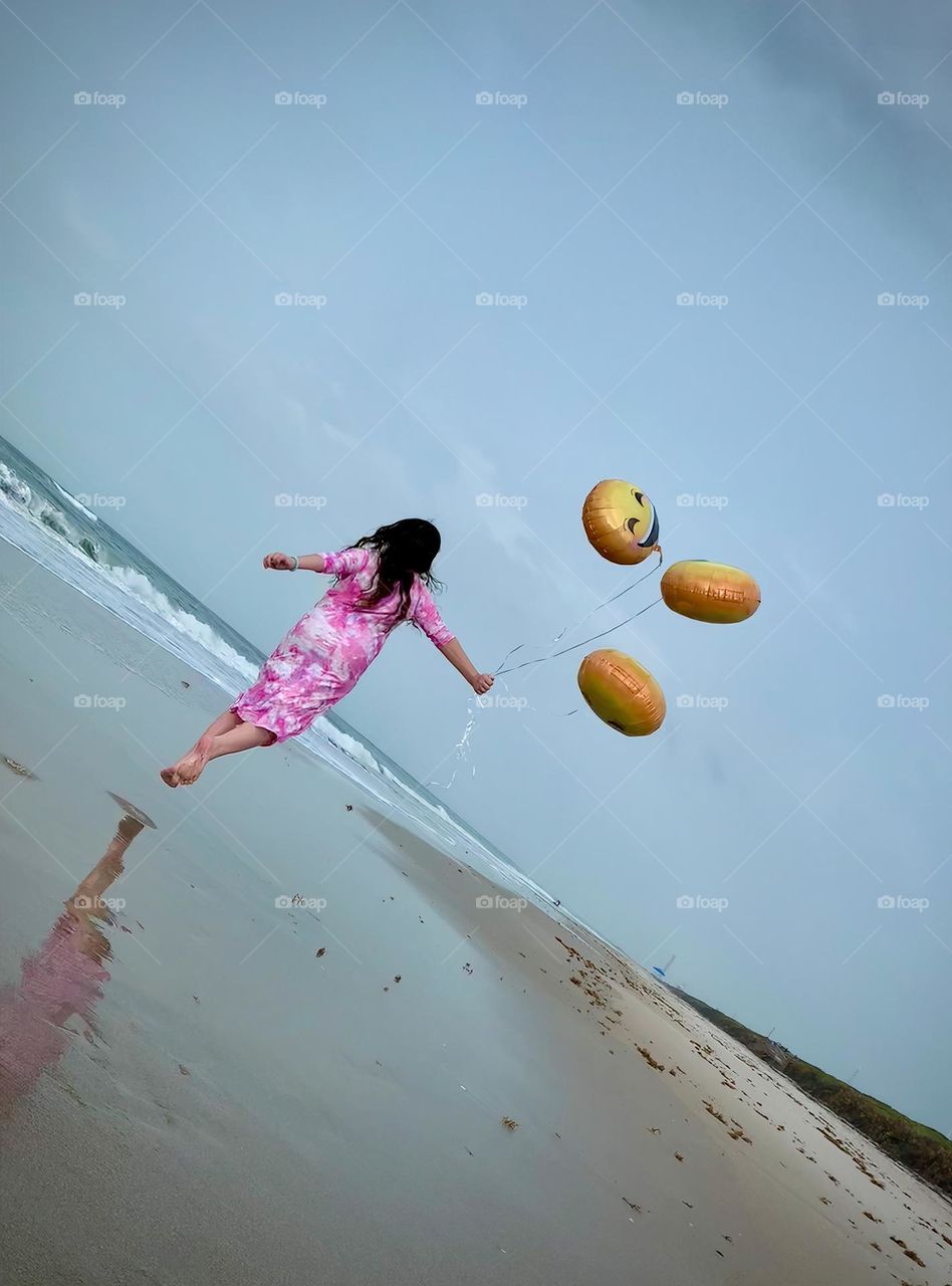 Little Girl Seems To Be Flying And Carried Up By The Three Helium Emoji’s Balloons On The Beach By The Ocean.