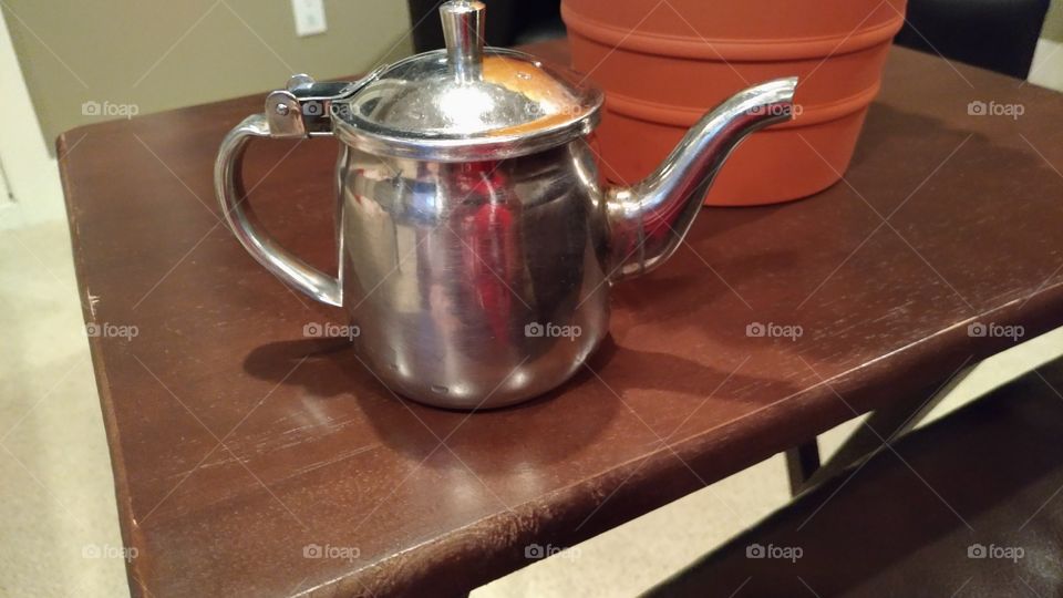 A tea pot, on a table, next to a vase, with a flower in it. Inside, a house, at a jaunty angle.