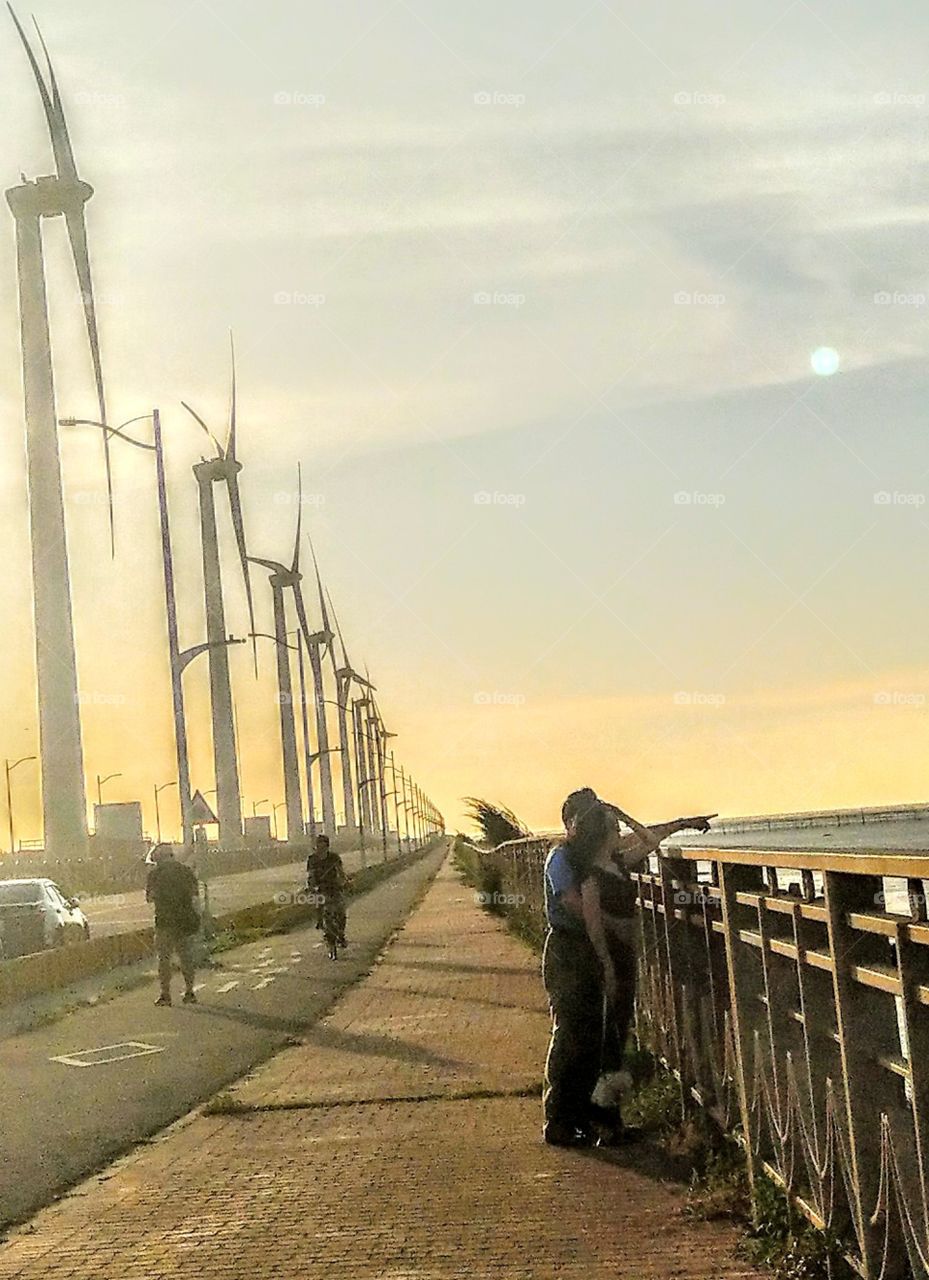 The art of composition: seaside and windmills landscape at dusk. backlit road. persons not very clear, but long road, sun light, windmill, people's action all were beautiful.