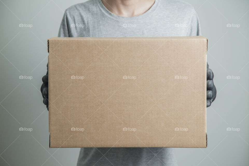 Detail of man with black gloves holding a cardboard box for delivering products on gray background. Delivery concept. Delivery service concept. Copy space.