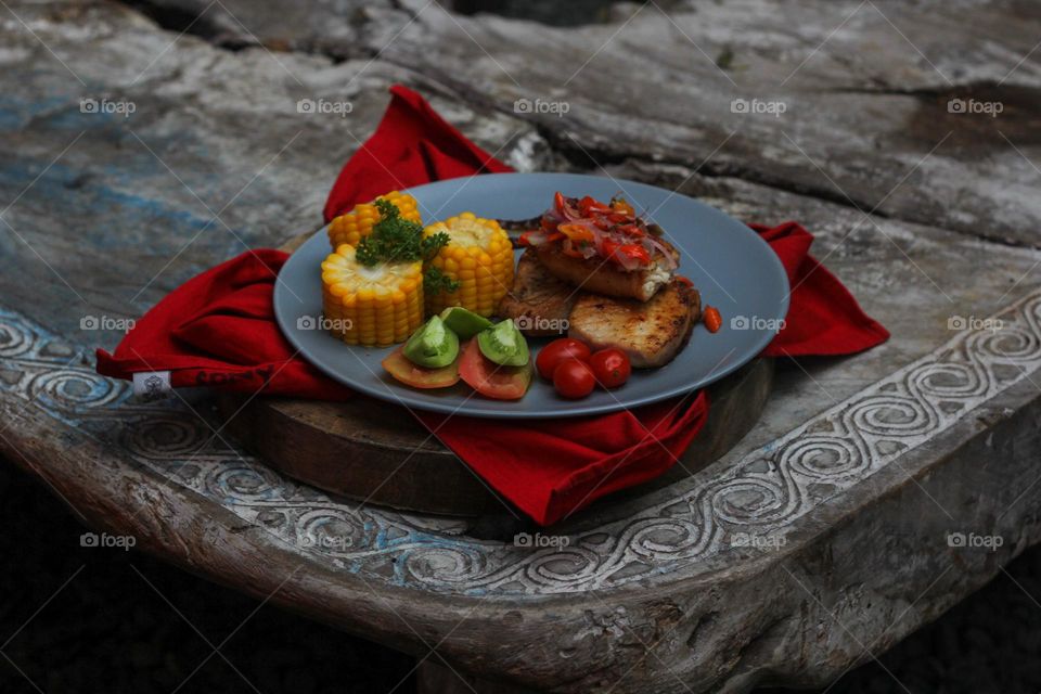 Tuna steak cooked with corn garnished with cherries, sliced tomatoes and lemon topped with sliced hot chili peppers on a baby blue melamine plate lined with red cloth and thick wood. Restaurant food.