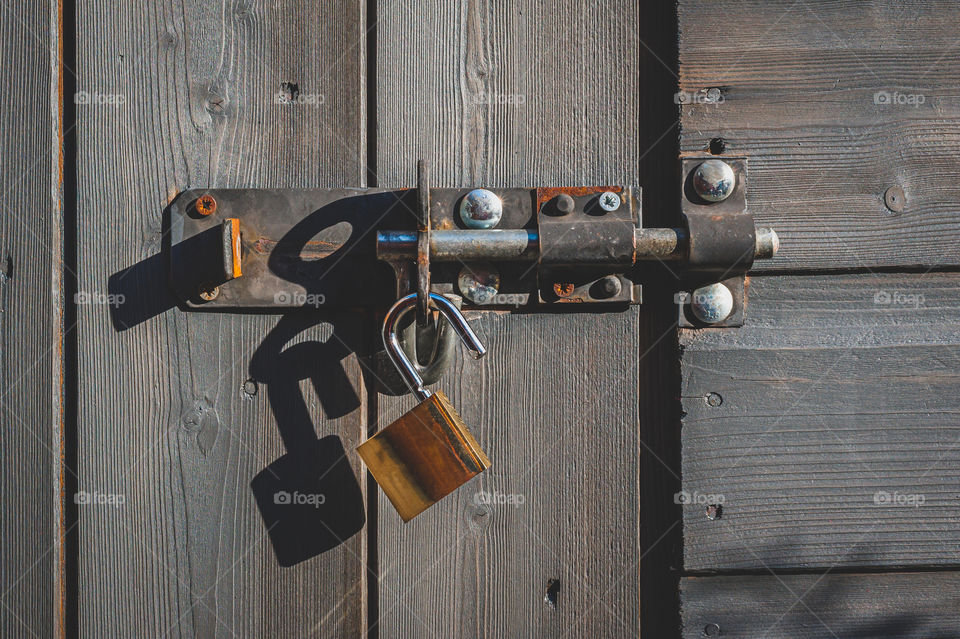 New shiny brass padlock hanging from old bolt lock on wooden door.