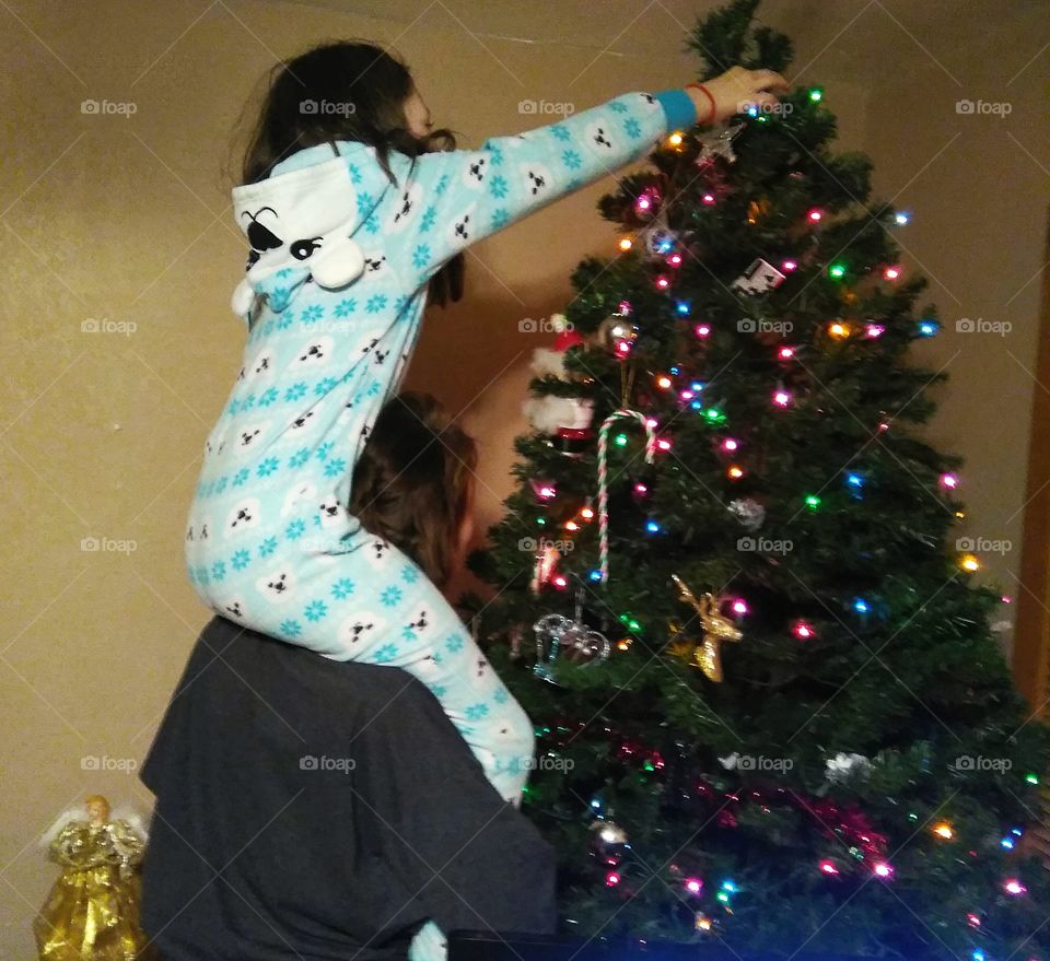 Molly (8) year old girl in her polar bear jammies on her momma's shoulders hanging ornaments high up on the colorfully lit Christmas tree.