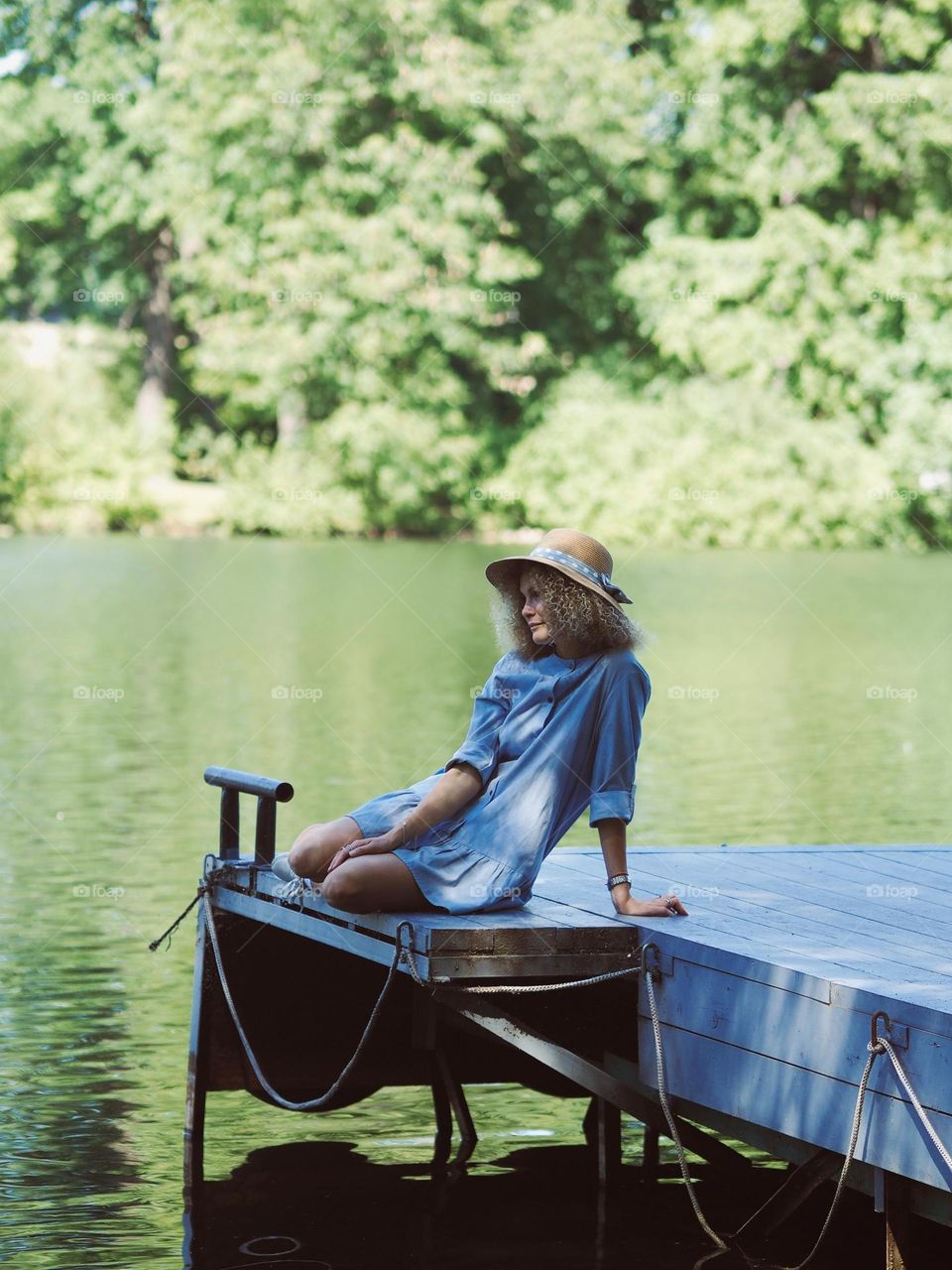 Young beautiful woman with curly hair wearing blue dress and hat sitting near water in park in sunny summer day, portrait of woman 