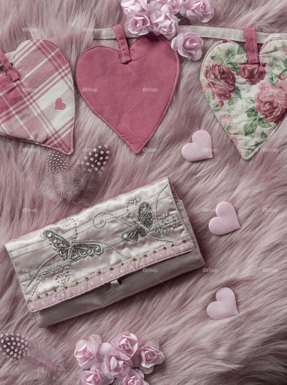 Pink hearts, paper flowers and feathers surround a silken purse on a pink faux fur background 