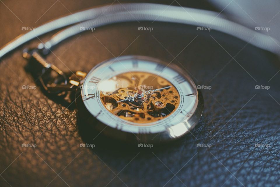 An intricate pocket watch showing roman numerals and a complex gear system behind the hands resting on a leather piece of furniture