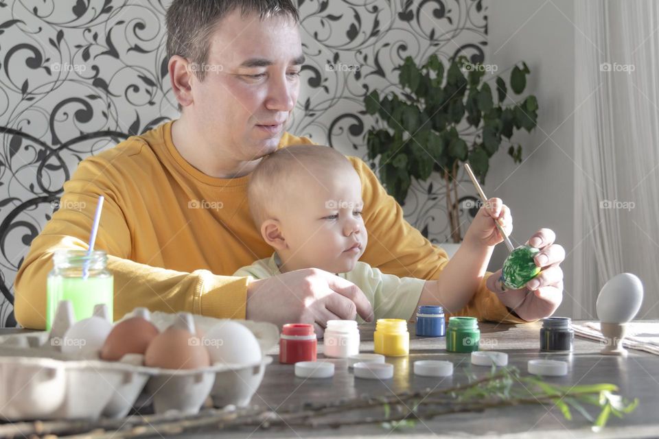 A little son and his father together in their free time paint Easter, chicken eggs with paints and brushes for the holiday. A child and a man create a craft and natural product in their family business.
