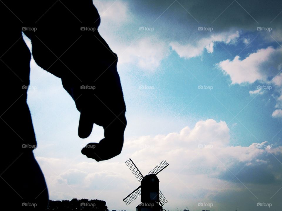 Silhouette of young boys arm and hand with a windmill in the distance on a blue moody cloudy sky
