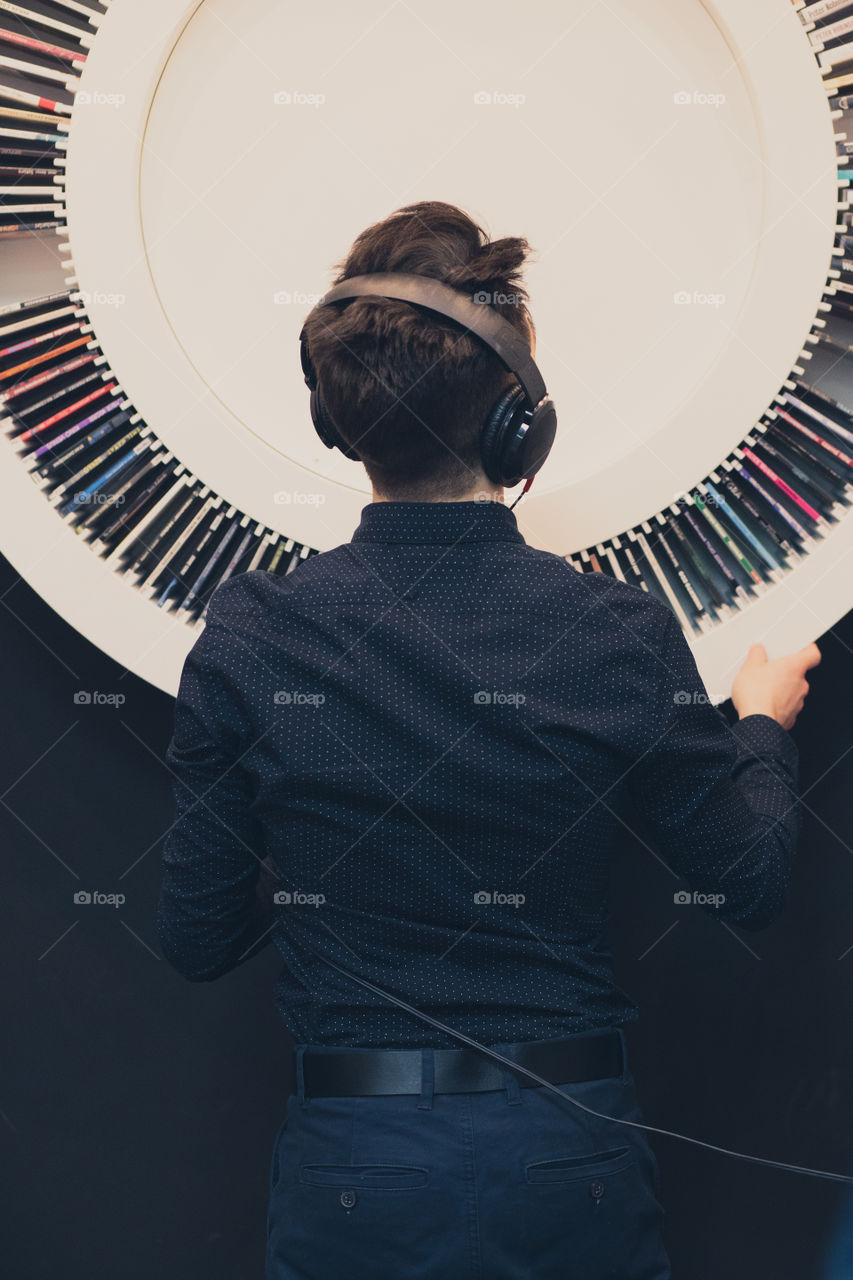 Young man listening to music through headphones standing next to bookshelf with recordings