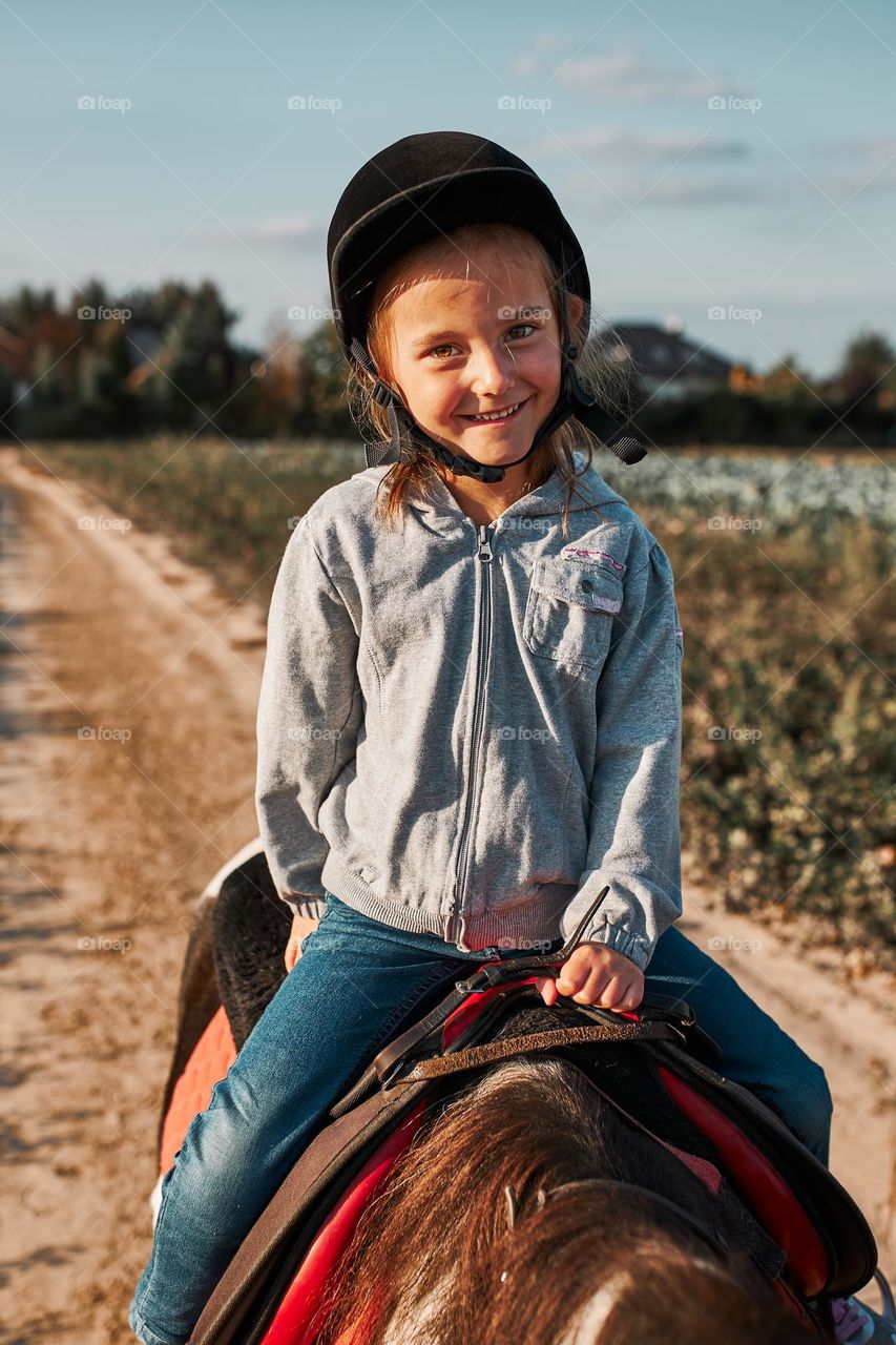 Little smiling girl learning horseback riding. 5-6 years old equestrian in helmet having fun riding a horse