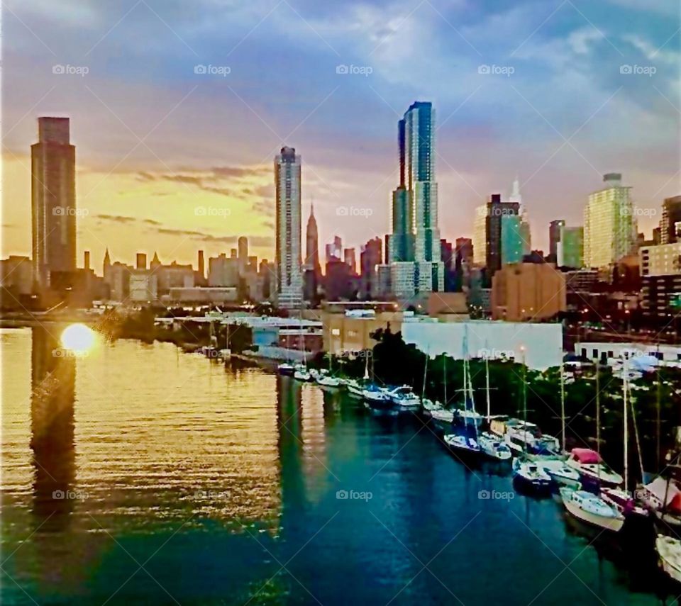 This is lovely “Newtown Creek” with a great variety of boats seen from the “Pulaski Bridge” in LIC, Queens at the golden hour of sunset in approximately 2020. The skyline in the far distance is “Manhattan”. Hypnotic Productions