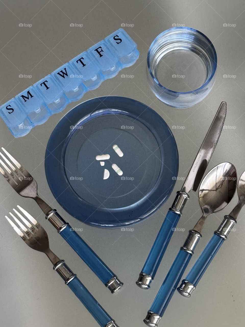 blue pink box with meds on plate with blue handle flatware and glass of water on frosted glass 