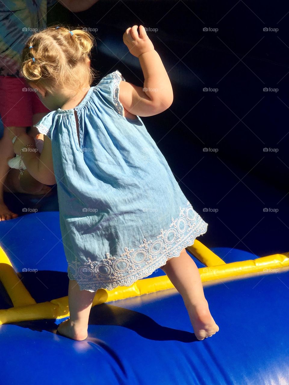 Unidentifiable blonde haired little girl toddler in baby blue lace frock and pigtails playing in outdoor playground, in summertime, from behind, copy space