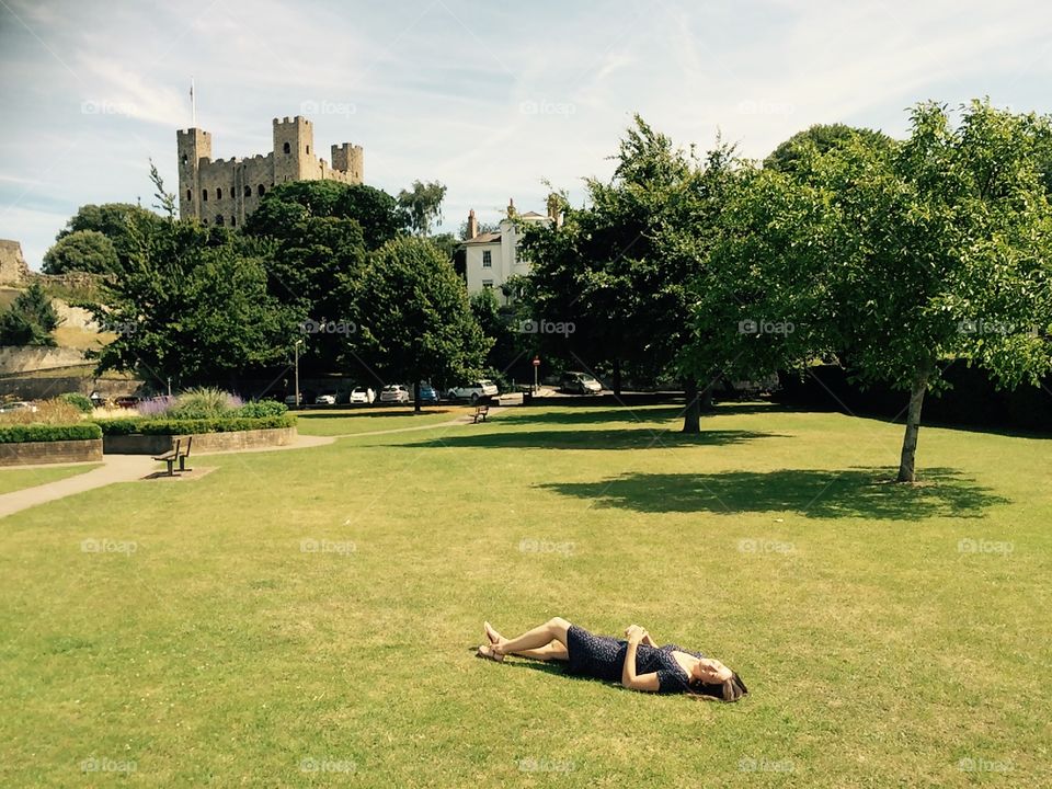 Young woman relaxing in a green park in Rochester, Kent overlooked by Rochester Castle. So laid back she’s horizontal! Taking it easy. Sit back and relax. Lying in a park. Sunbathing. English Park. 