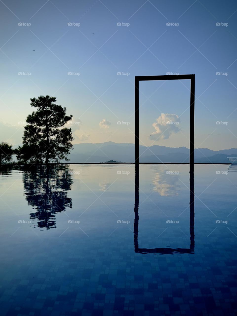 8:13 - At sunset, an infinity pool stands alongside a stunning tree and a rectangular shaped rack, while the blue sky dotted with fluffy white clouds fills the air with a summertime ambiance