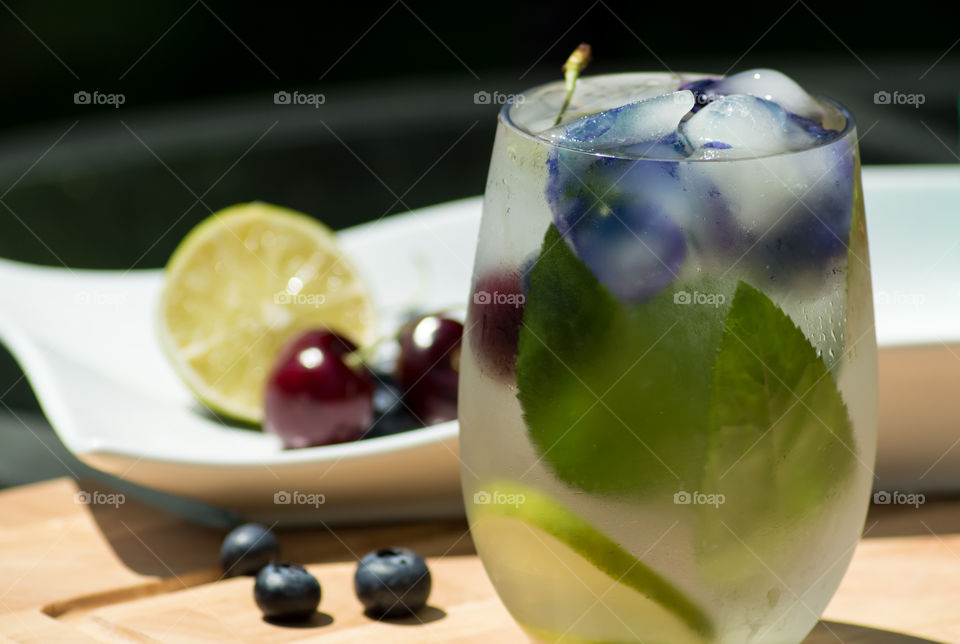 Beautiful gourmet summertime drinks background with cold frosty glass of lime juice, cherry and mint flavored water garnished with edible flower ice cubes on wood background outdoors 
