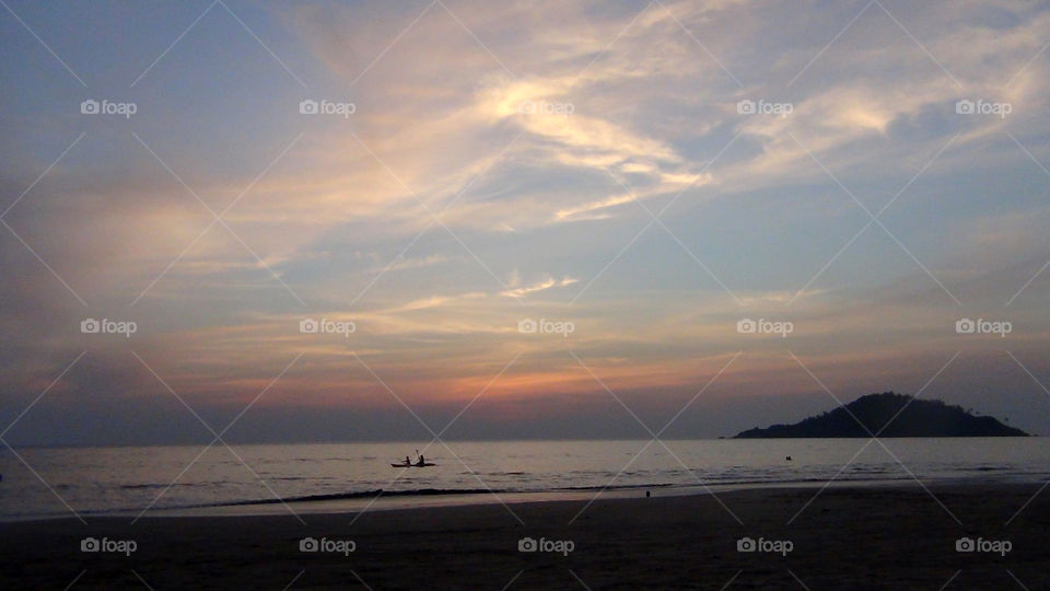 A calm sea with a colourful pleasant sunset, silhouette of a couple kayaking and a small island.