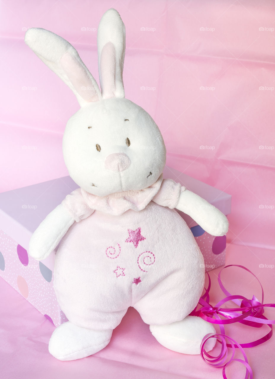 A cuddly soft pink toy rabbit standing beside a spotted giftbox.