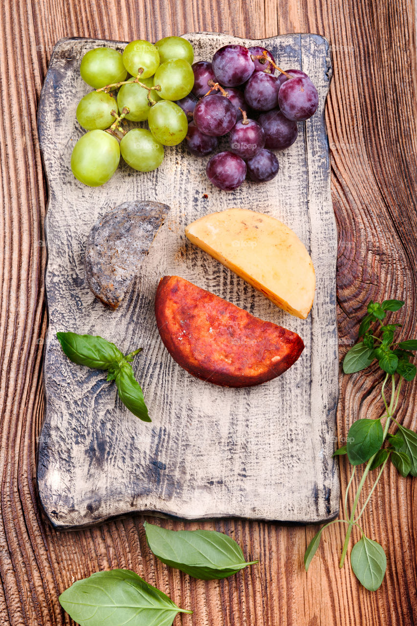 Cheese and grapes decorated with basil leaves on handmade pottery plate on old wooden table from above