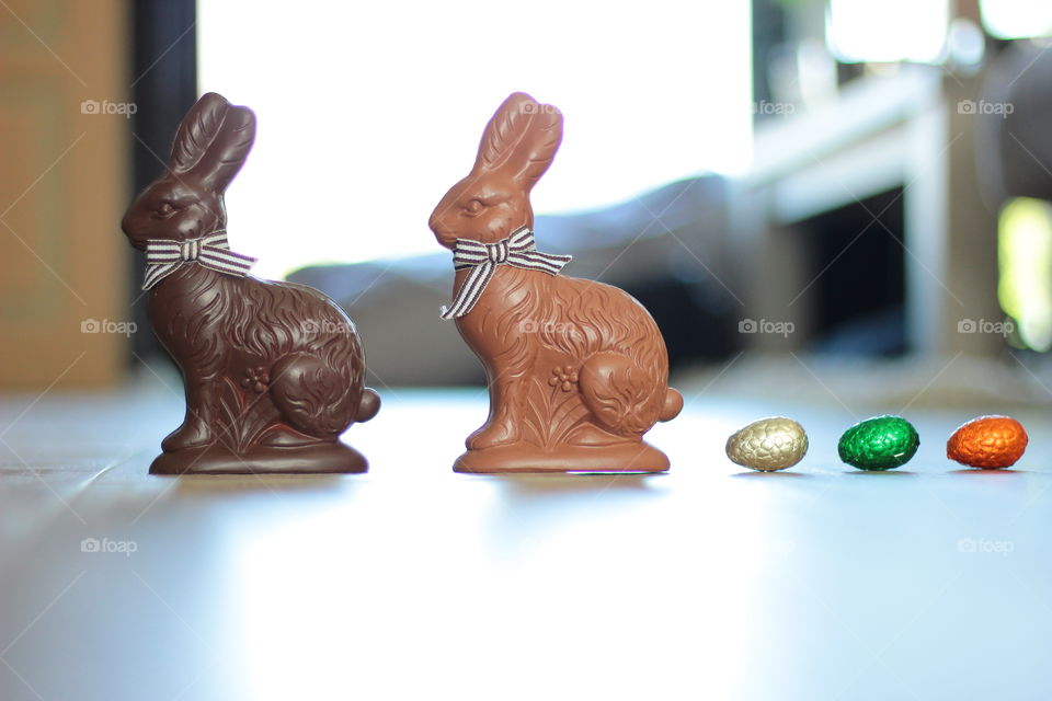 A portrait of two chocolate easter bunnies with a row of chocolate eggs behind them. each one of the eggs is wrapped in colorful tin foil.