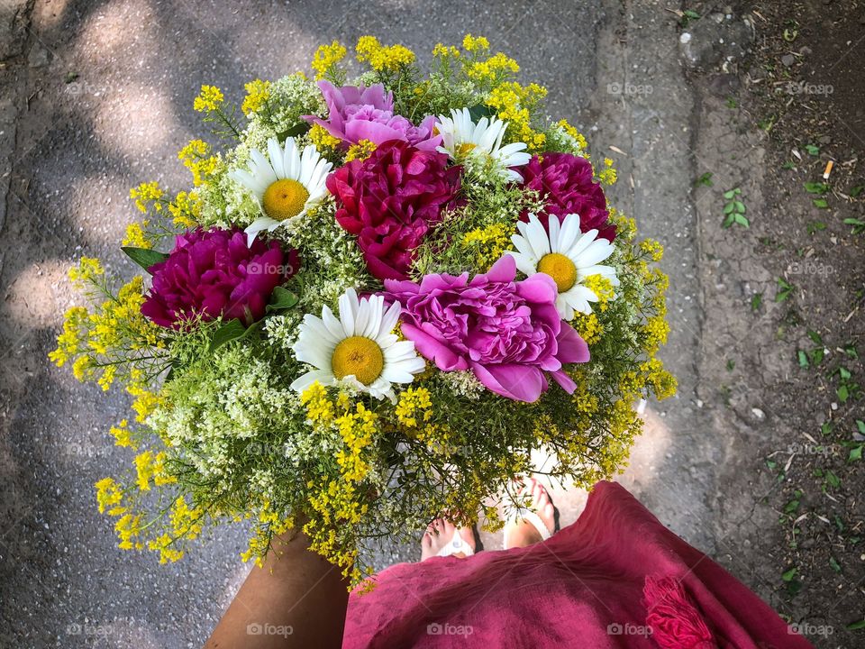 Woman in pink dress holding a giant bouquet of summer flowers consisting of pink peonies, daisies and yellow goldenrod