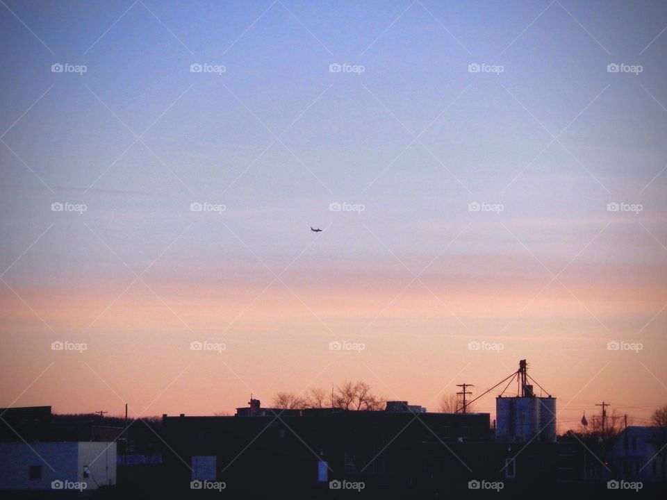 Airplane flies over small rual town in Michigan as the sun sets turning the sky and clouds pink and orange