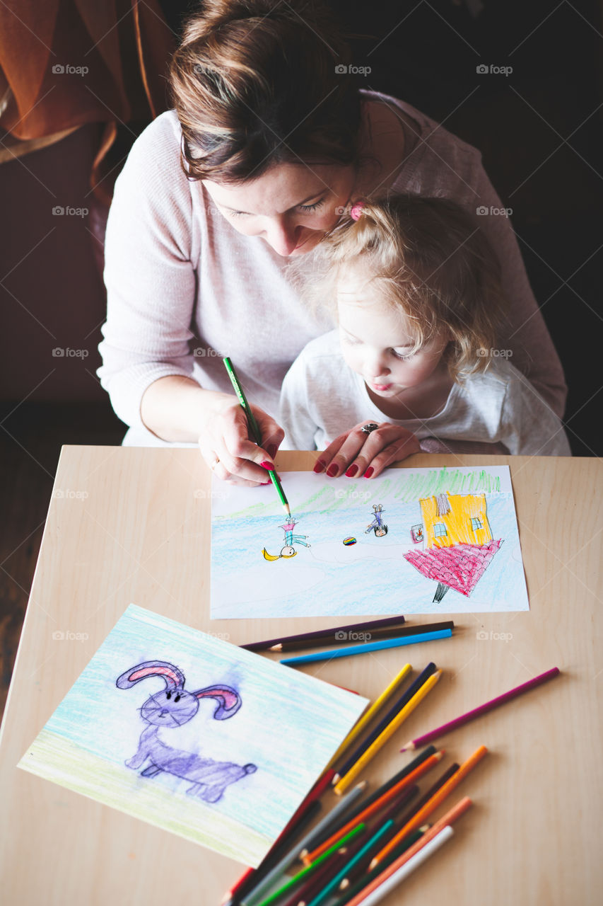 Mom with little daughter drawing a colorful pictures of house and playing children using pencil crayons sitting at table indoors. Shot from above