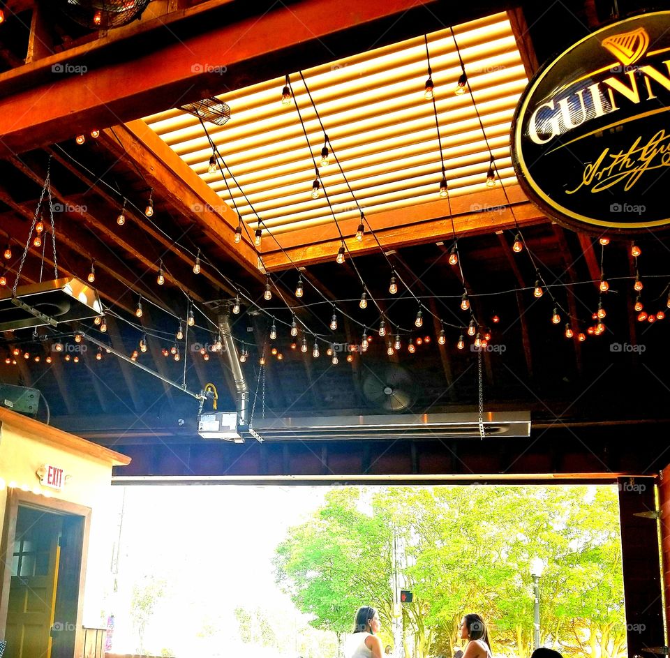 Summer day views from the neighborhood pub patio overlooking bright green trees with glowing gold string bulb lights draped across louvered open air ceiling with classic Guinness beer sign.