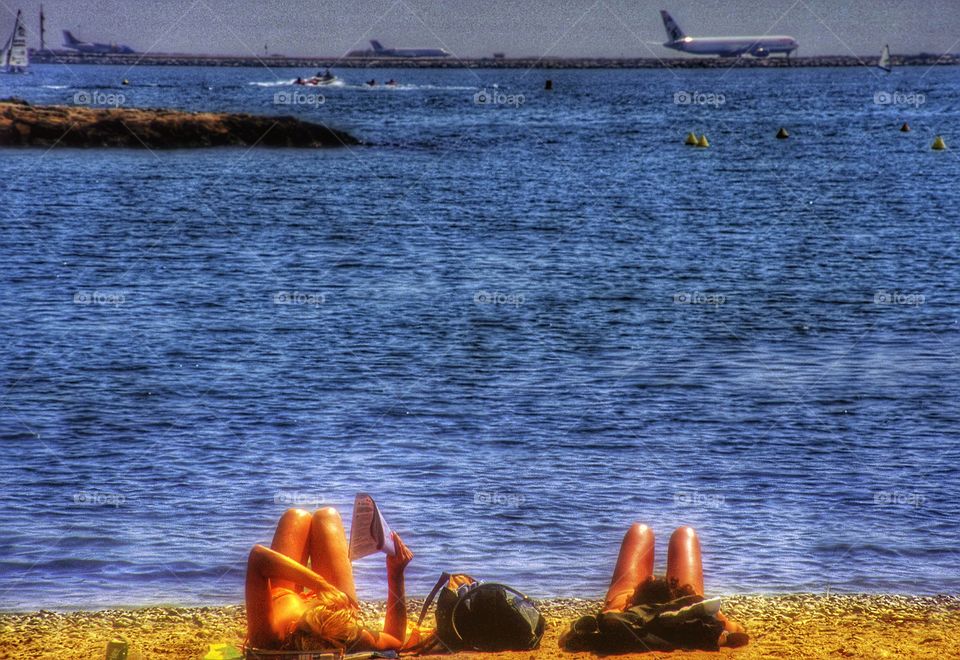 Beach. Two women sunbathing on beach airport with jet taking off in distance 