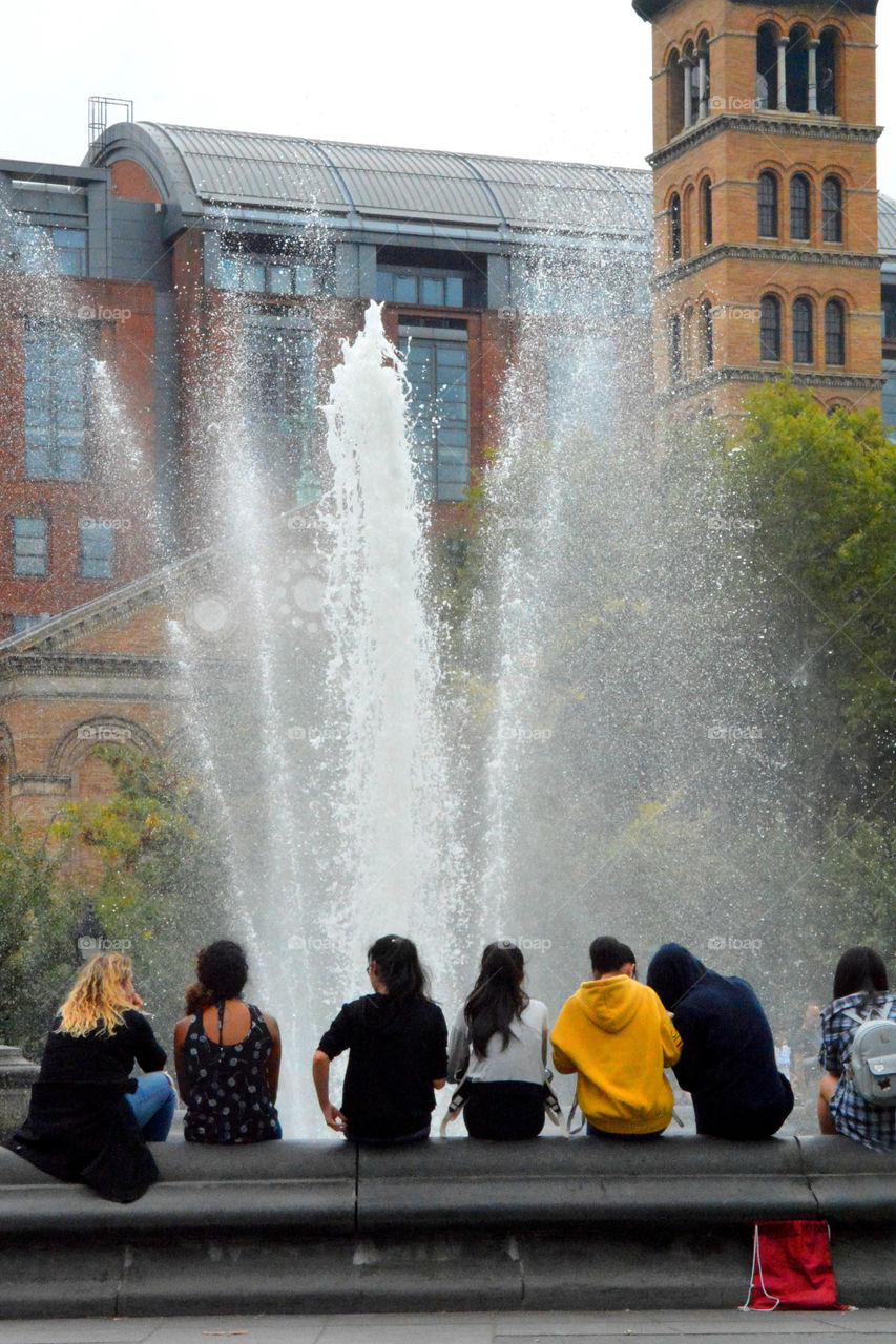 Unrecognizable people with their backs towards the camera sitting around a fountain in Washington Square park New York City.