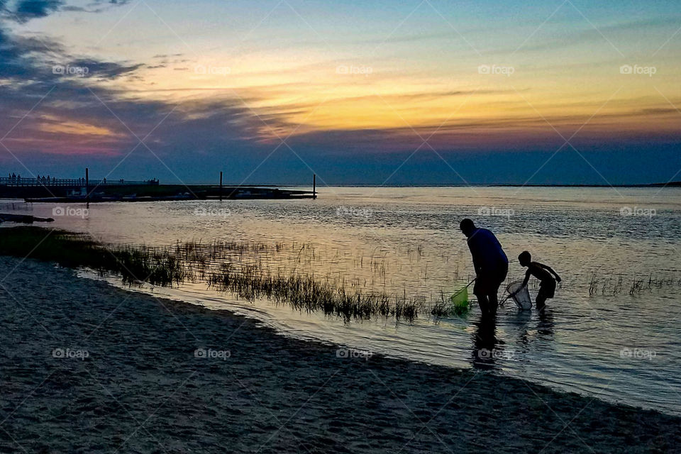 Silhouettes of father and son wading in the water on a beach at sunset.