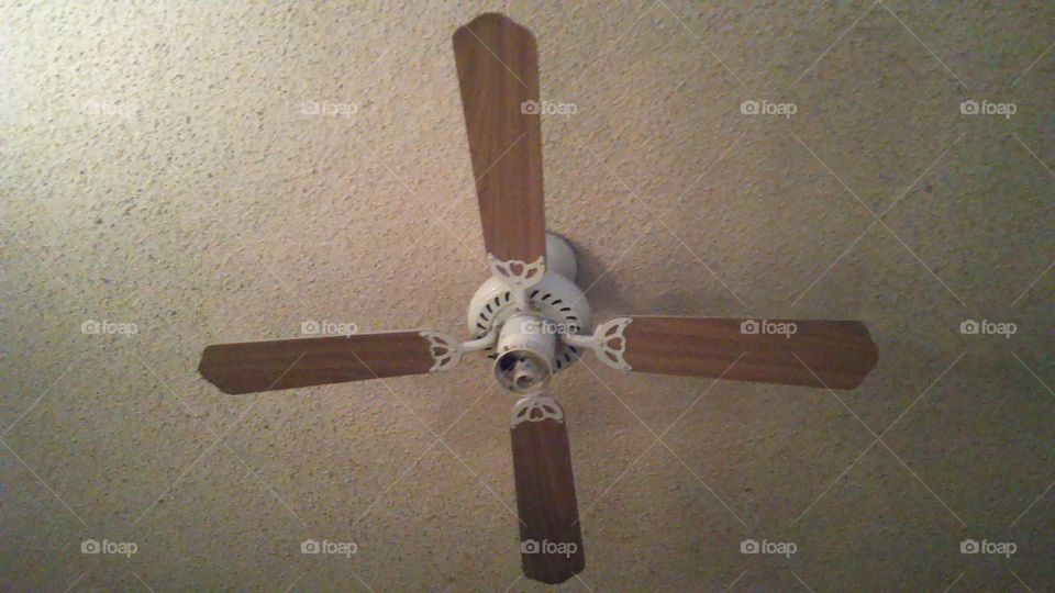 The symmetrical blades of a ceiling fan against a 1970's "popcorn" style acoustic ceiling treatment.
